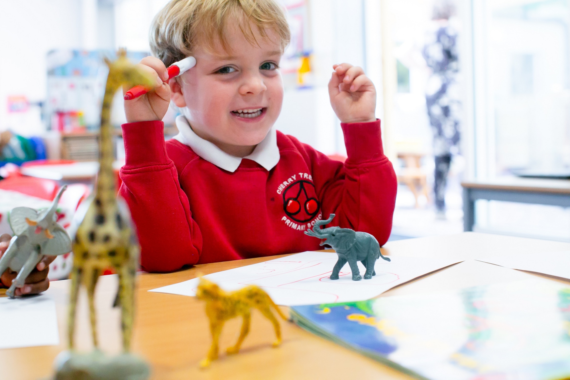 a pupil holds a pen and smiles. There are small plastic dinosaurs on the desk